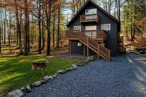A cabin with a wooden staircase leading to a balcony, surrounded by a forest and a deer on the grass. - The Spot Lake Wallenpaupack-WLE (Lake Ariel)