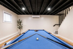A pool table with a blue felt surface, surrounded by white walls, a potted plant, and a staircase with black railings. - The Spot Lake Wallenpaupack-WLE (Lake Ariel)