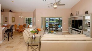 A spacious living room with a sofa, a glass coffee table, and a TV. There is a dining area with chairs and a table, and a kitchen area with a counter and cabinets. - Alecia -Gulf access Home (Cape Coral)
