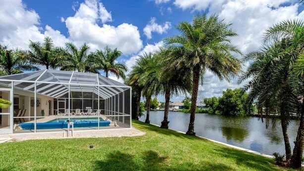 A covered swimming pool with a glass enclosure, surrounded by palm trees and a body of water. - Alecia -Gulf access Home (Cape Coral)