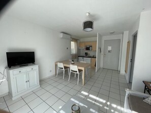 A modern living room with a flat-screen TV, a dining table with chairs, a kitchen area with cabinets and appliances, and a door leading to another room.