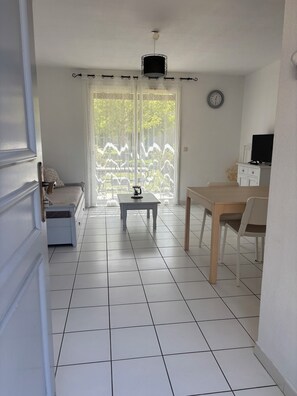 A room with a dining table, chairs, a small table with a coffee maker, a sofa, and a window with curtains.