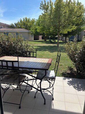 An outdoor patio with a tiled table and chairs, surrounded by greenery and a house in the background.