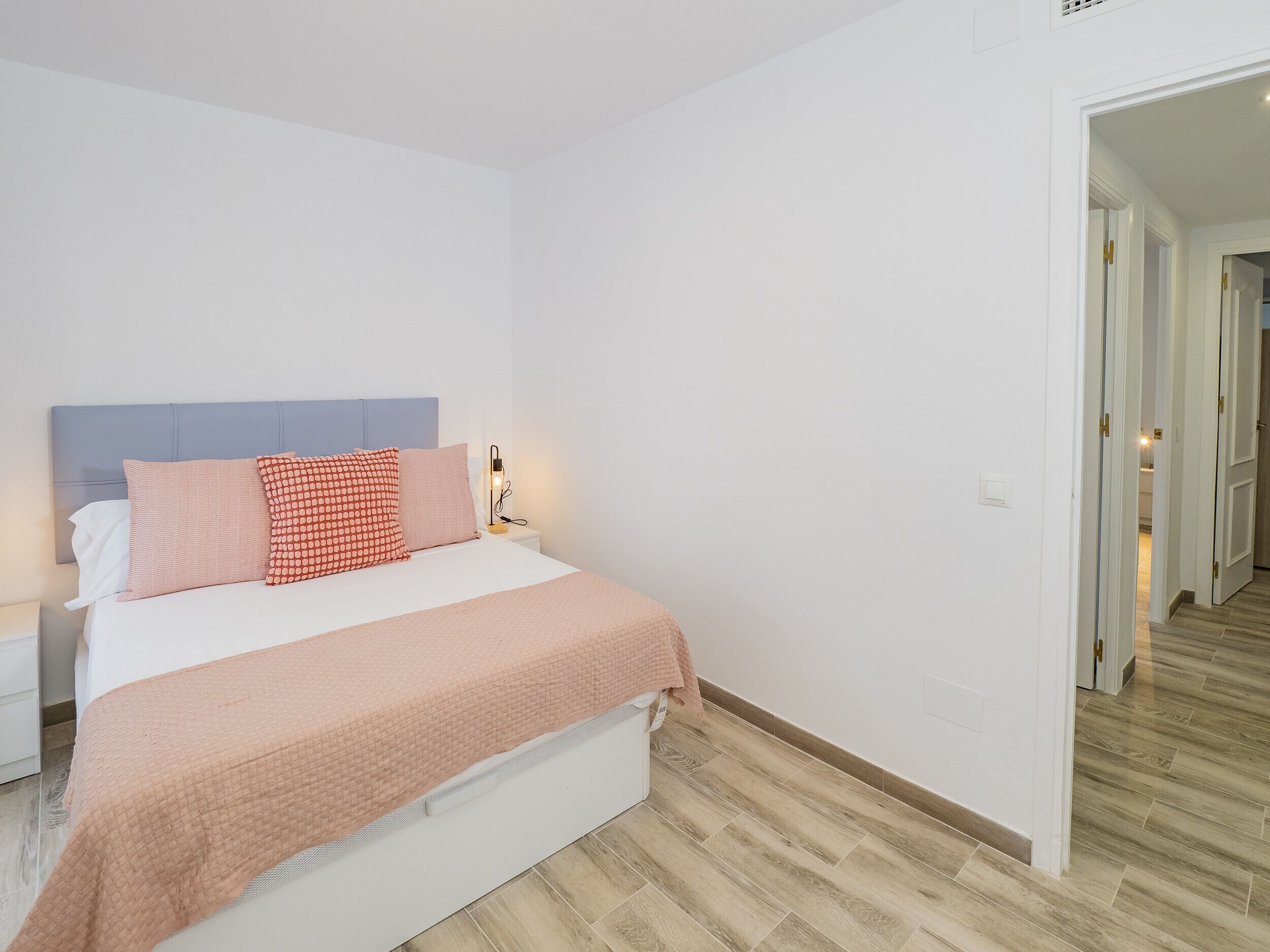 A neatly made bed with a headboard, a bedside table with a lamp, and a wooden floor.