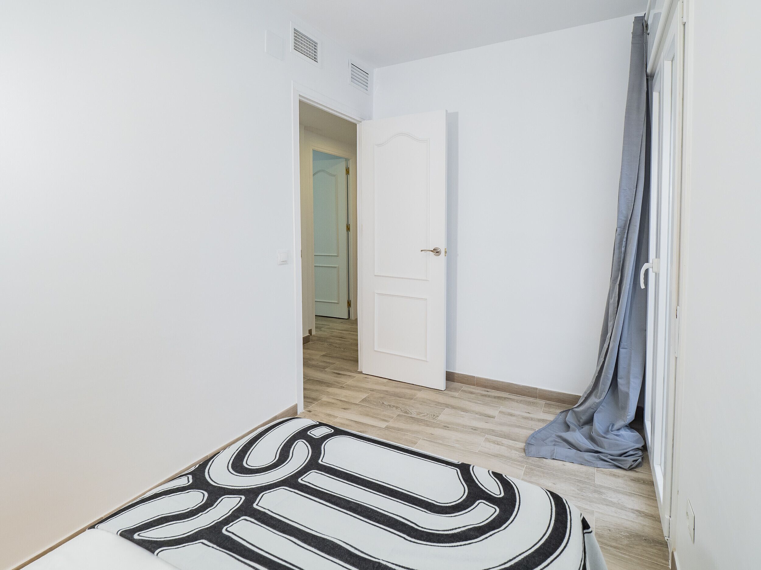 A narrow hallway with a white door at the end, a grey curtain partially covering a window, and a patterned rug on the floor.