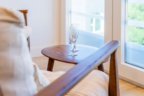 A wooden chair with a cushion, a small round table with a clear glass on it, and a window with a view of greenery.