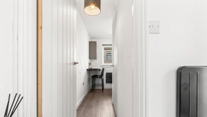 A narrow hallway with white walls and wooden flooring, leading to a kitchen area with a window and a chair. - City Centre House with EV Parking  (Coventry)