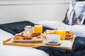Bandeja de desayuno con tortitas, huevos, tocino, tostadas y zumo de naranja sobre una cama con una manta y almohadas oscuras.