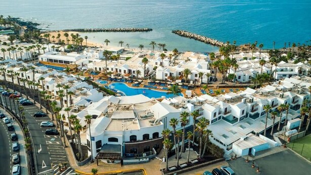 An aerial view of a coastal resort with a central swimming pool, white buildings, and palm trees.
