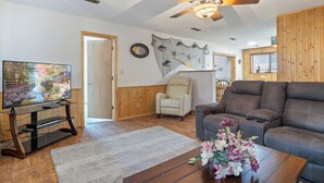 A living room with a television, a sofa, a coffee table with flowers, a ceiling fan, and a wall decorated with marine life.