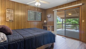 A bedroom with a bed, a ceiling fan, a window with blinds, a desk with a chair, and a sliding glass door leading to an outdoor area.