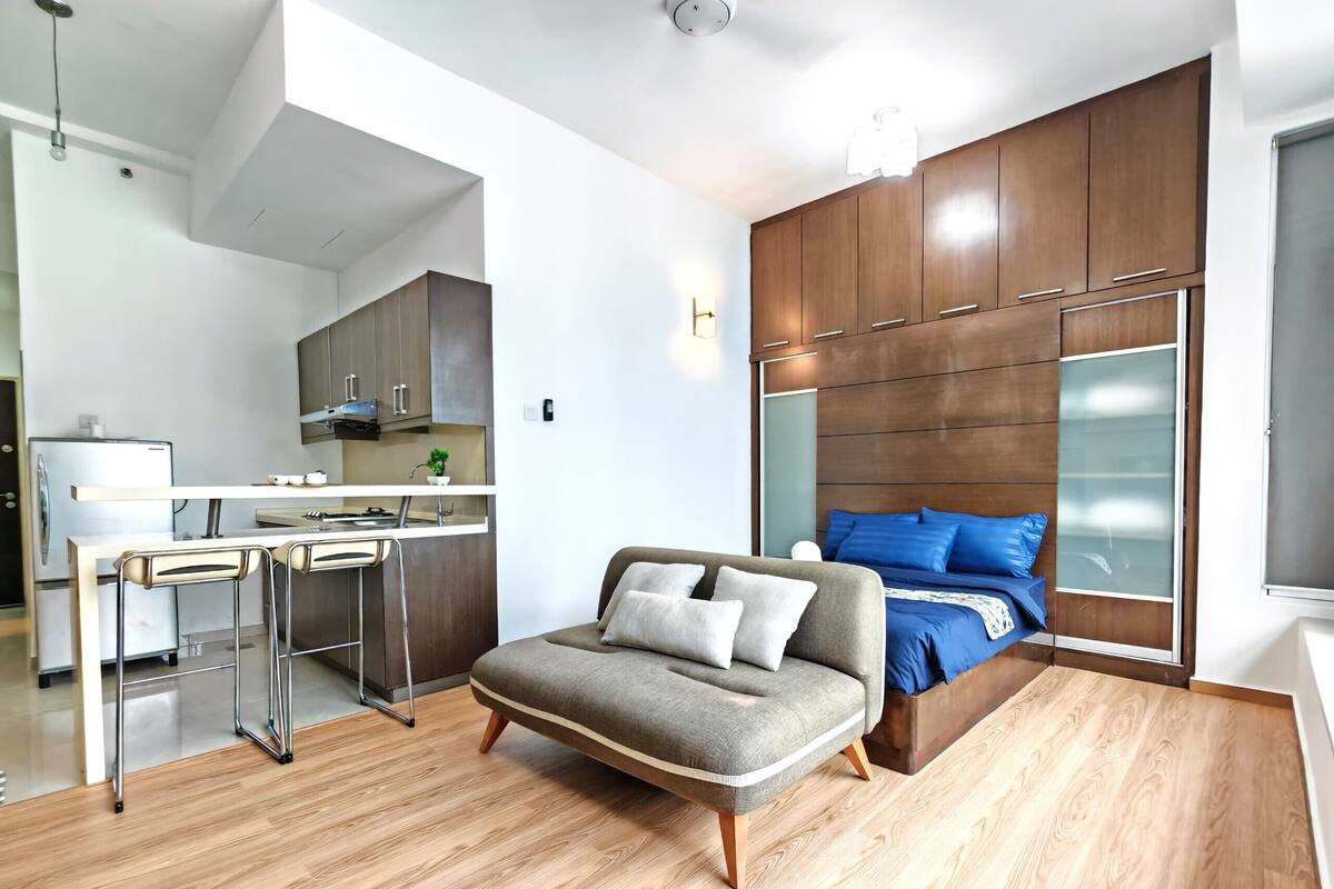 A modern apartment with a kitchenette, a sofa, a bed with blue bedding, and wooden flooring.