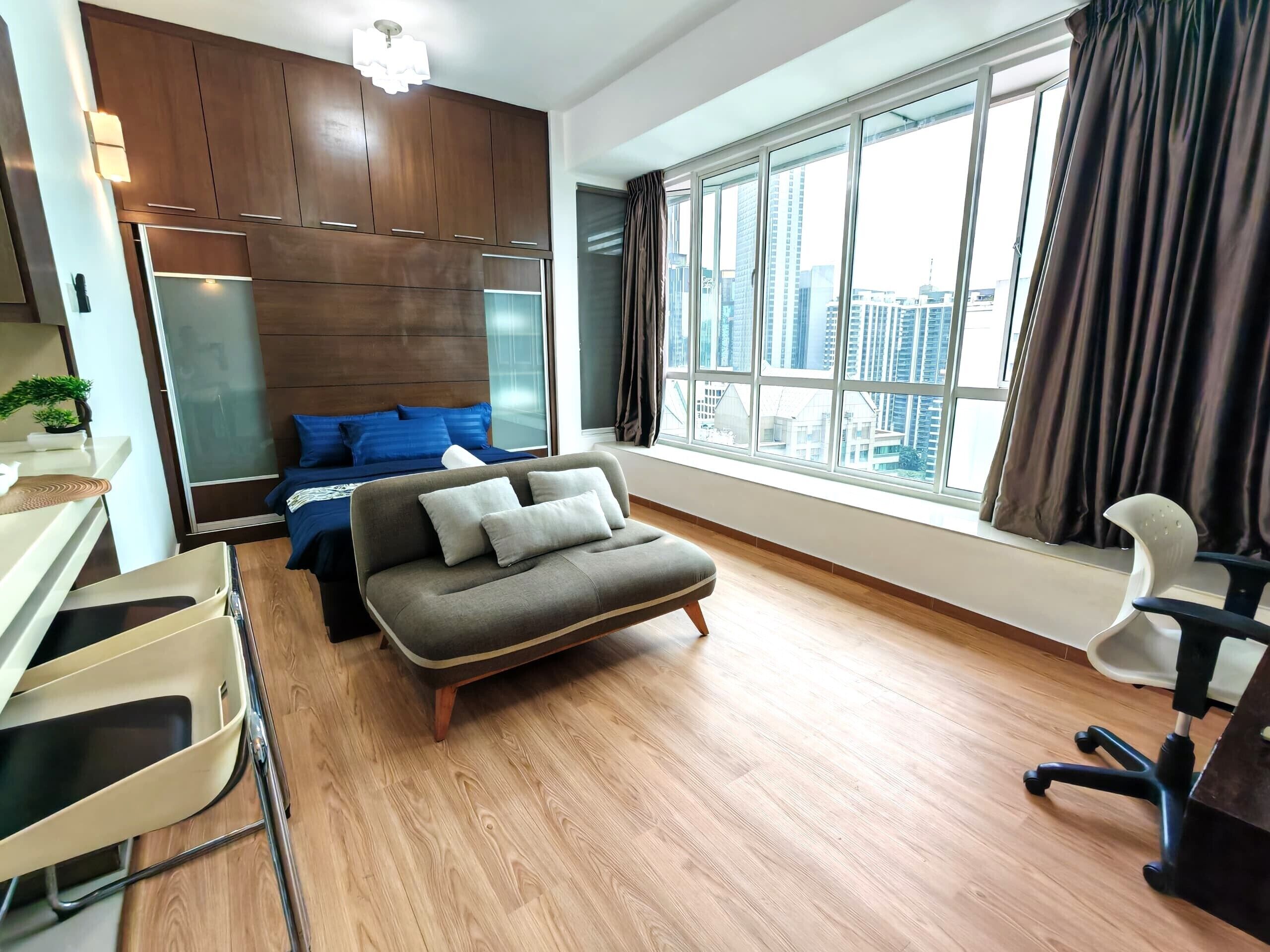 Une chambre d’hôtel moderne avec un plancher en bois, un canapé, un bureau avec une chaise, un lit avec une literie bleue et de grandes fenêtres avec des rideaux.
