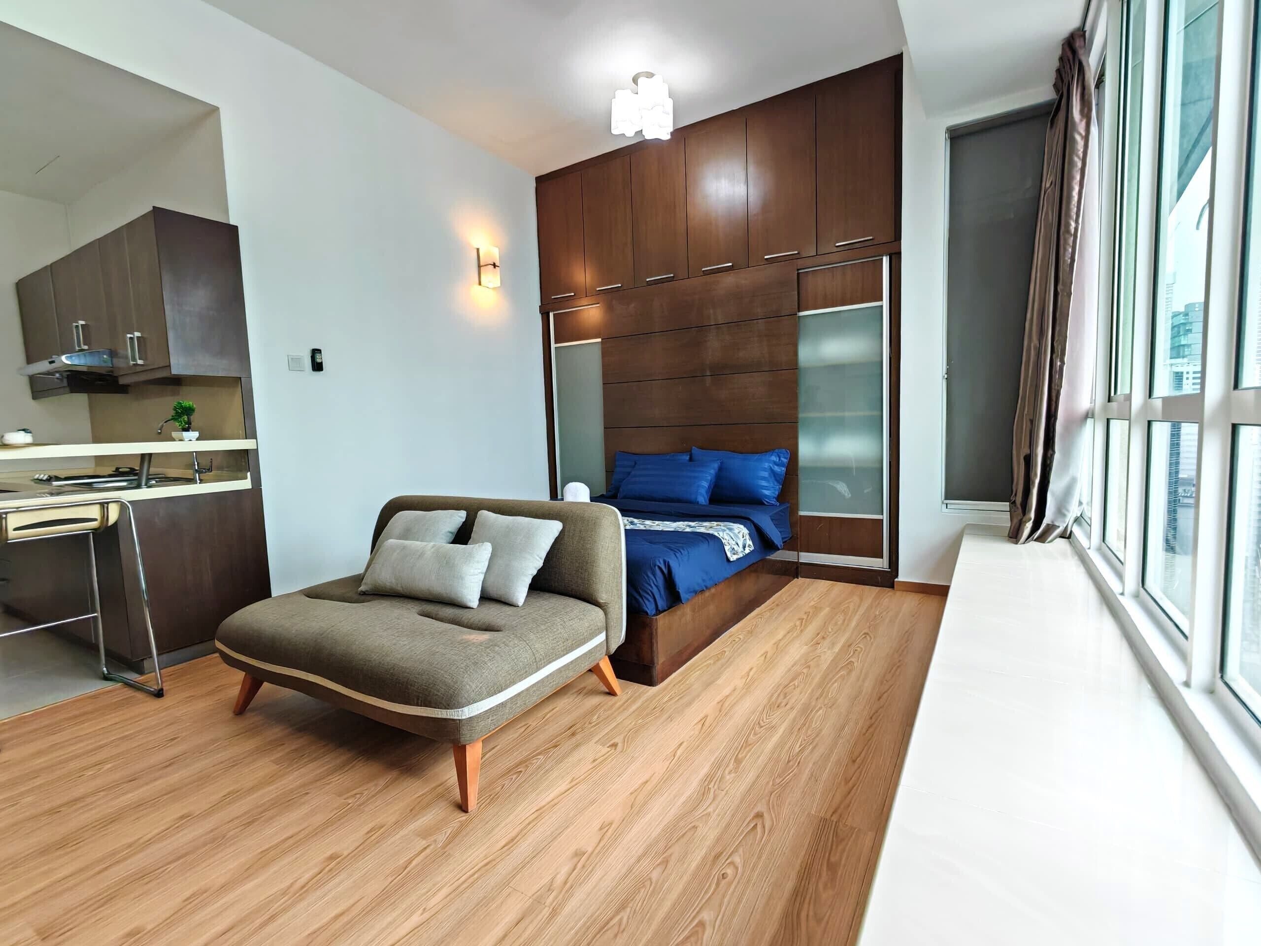 A modern bedroom with a wooden bed, a sofa, a built-in wardrobe, and a kitchen area.