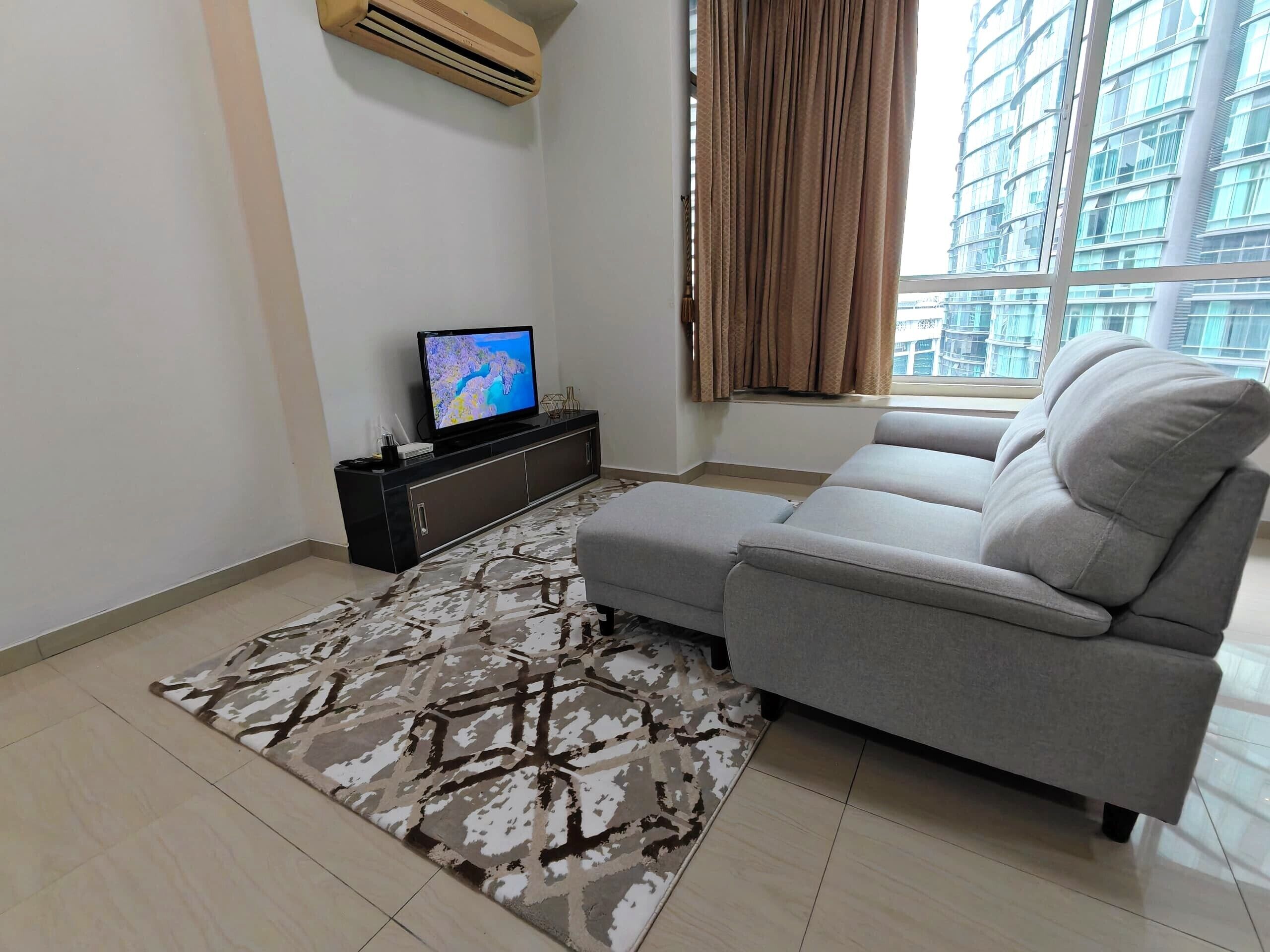 A modern living room with a grey sofa, a patterned rug, a flat-screen TV on a black entertainment unit, and a large window with curtains.