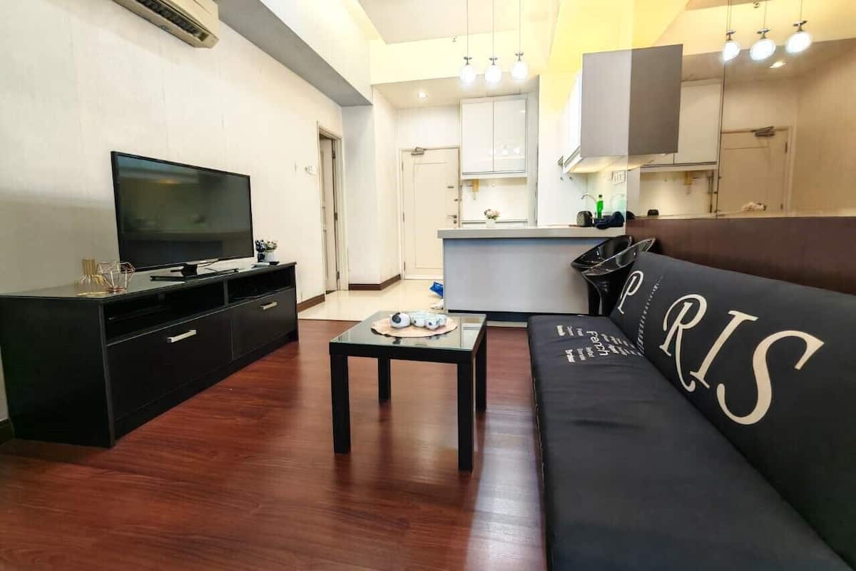 A modern living room with a black sofa, a flat-screen TV, a coffee table, and a kitchen area in the background.