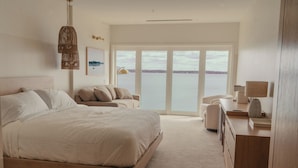 A bedroom with a large bed, a sofa, a desk with a lamp, and a view of the water through large windows. - The Shoreline Hotel (Spring Park)