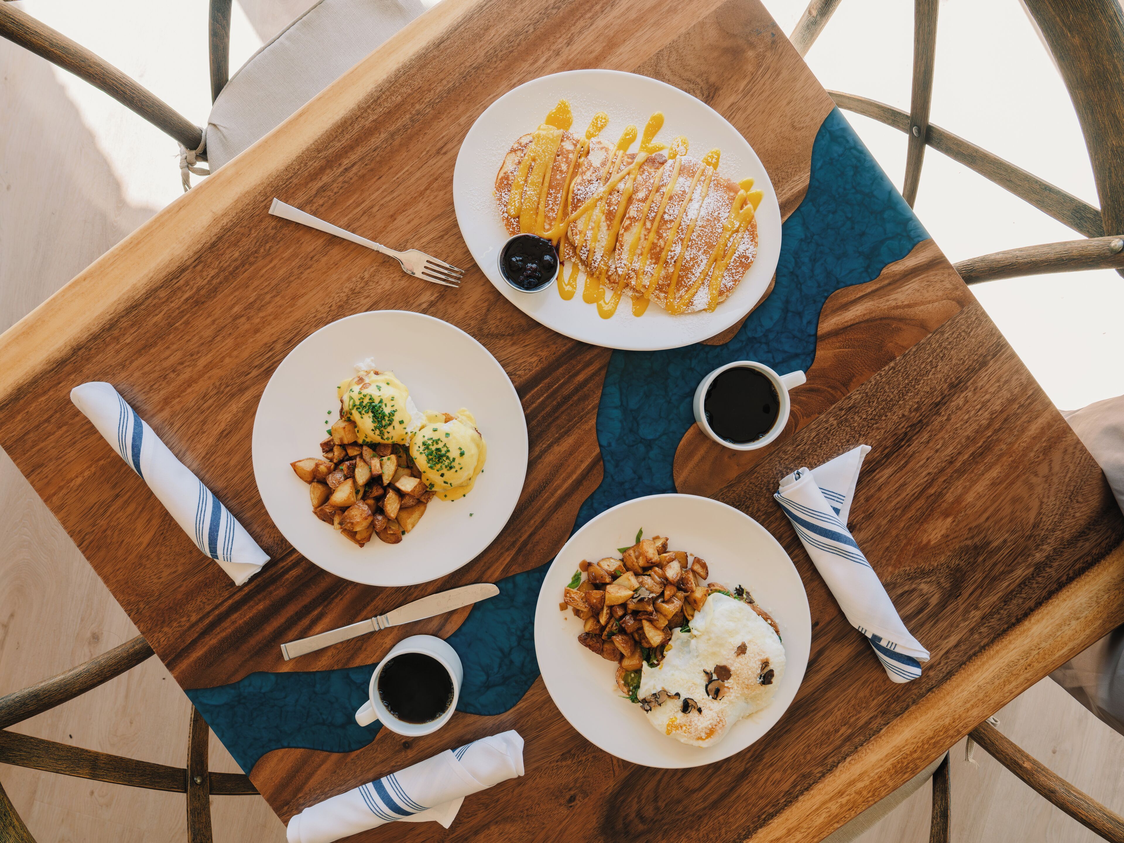 A wooden table with a blue placemat, holding plates of food, a fork, a knife, and two cups of coffee.