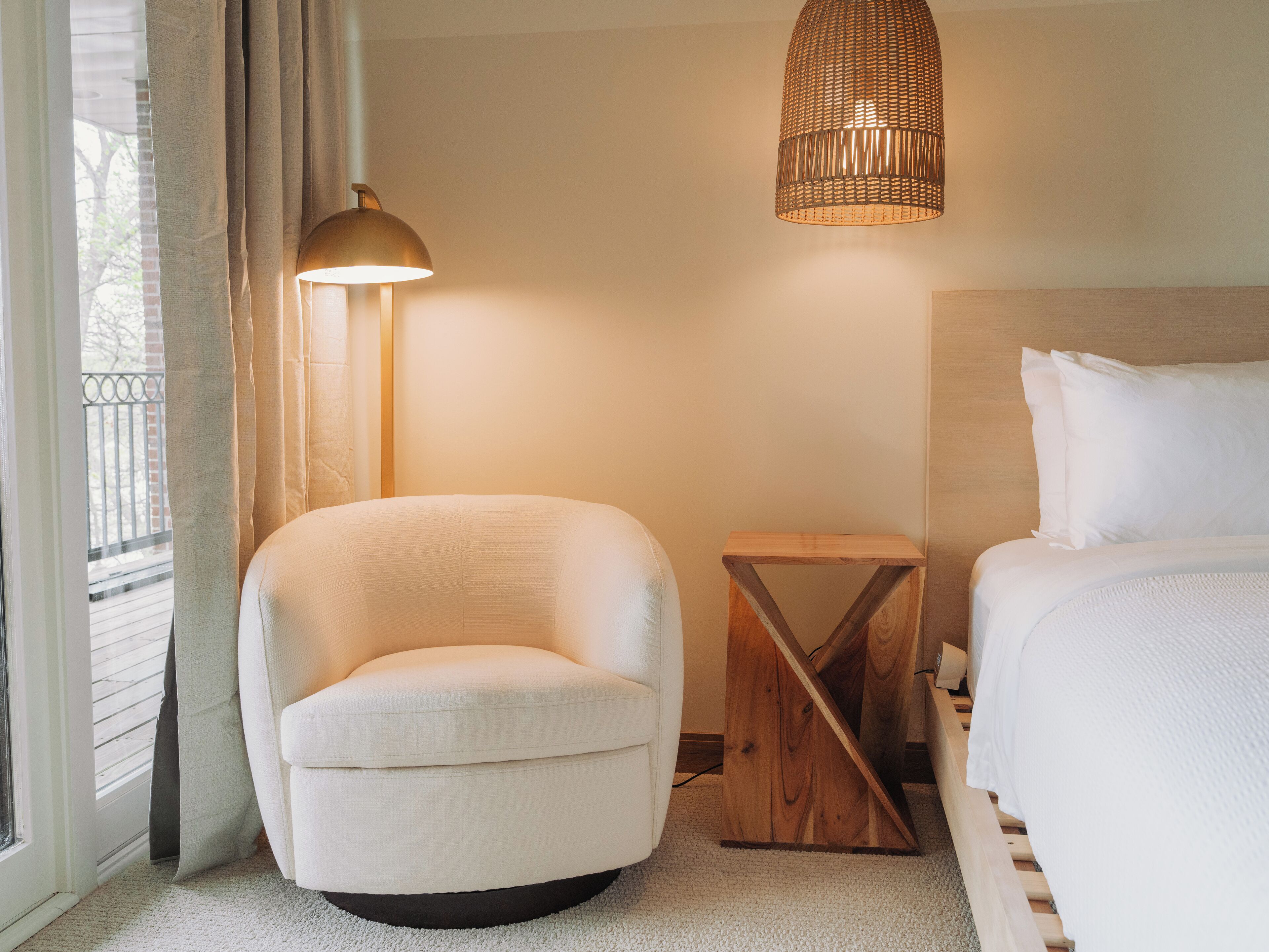 Ein Hotelzimmer mit einem Bett, einem Sessel, einem kleinen Holfnachtkästchen, einer Stehlampe und einem Fenster mit Vorhängen.