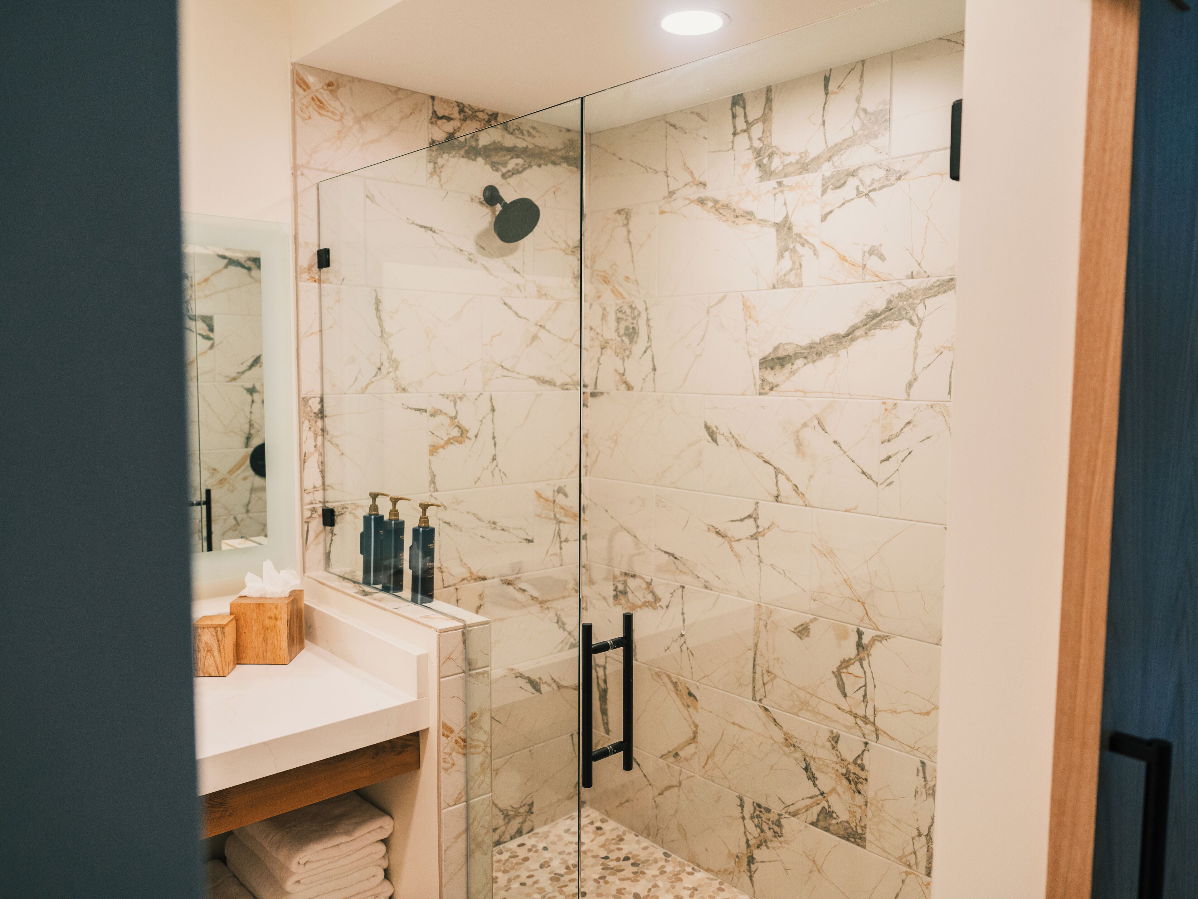 Un baño moderno con baldosas de mármol, cabina de ducha con mampara de vidrio y tocador de madera con artículos de aseo.