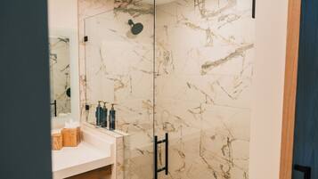 Un baño moderno con baldosas de mármol, cabina de ducha con mampara de vidrio y tocador de madera con artículos de aseo.