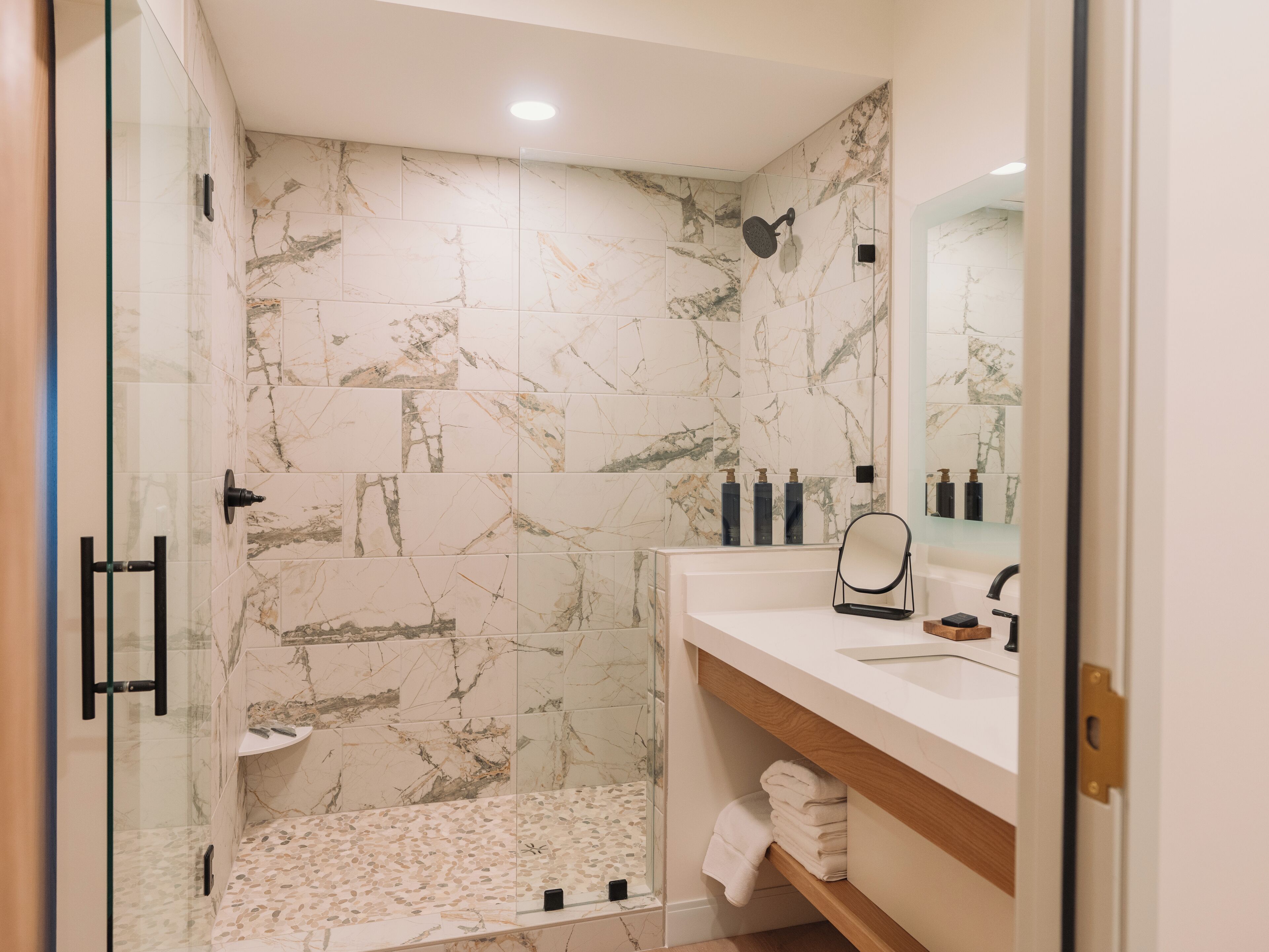 A modern bathroom with marble tiles, a glass shower enclosure, a white sink, and a wooden vanity.