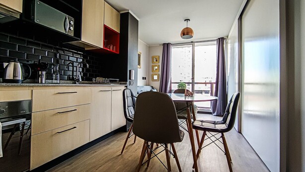 Een kleine keuken met moderne apparatuur, een eethoek met een ronde tafel en stoelen, en een groot raam met gordijnen.