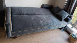 Een donkergrijze bank met gouden accenten, geplaatst tegen een muur met een raam en een klein fotolijstje.