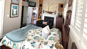 Un dormitorio con cama, lámpara de noche, ventilador de techo, chimenea y televisor.