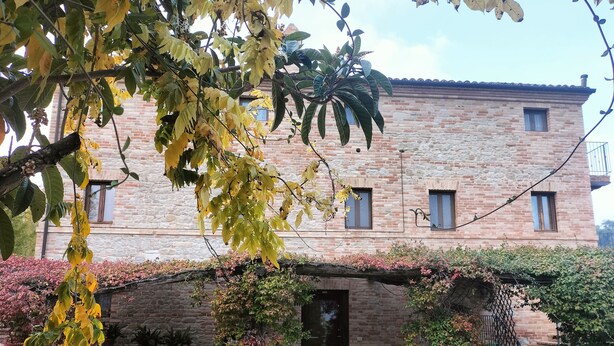 A stone building with a brick facade, a covered patio with outdoor seating, and a garden area with plants and shrubs. - Cottage on the Marche hills with swimming pool. (Marche)