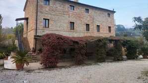 A two-story stone building with a balcony, surrounded by gravel and potted plants. - Cottage on the Marche hills with swimming pool. (Marche)