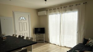 A modern living room with a sofa, a dining area with a table and chairs, a flat-screen TV on a stand, and large windows with curtains. - GITES BETWEEN SEA AND MOUNTAINS (Appietto)