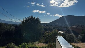 A clear sky with the sun shining through scattered clouds, a mountain in the background, and a residential area with houses and greenery. - GITES BETWEEN SEA AND MOUNTAINS (Appietto)