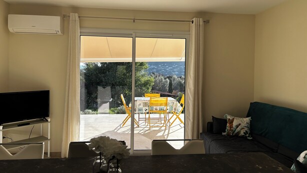 A living room with a TV, a sofa, a dining table with chairs, and a large sliding glass door leading to an outdoor patio with a yellow table and chairs. - GITES BETWEEN SEA AND MOUNTAINS (Appietto)