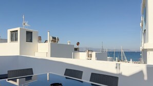 A modern rooftop terrace with a glass table and chairs, white walls, and a view of the sea and boats. - Naxian Downtown Residence (Naxos Chora)