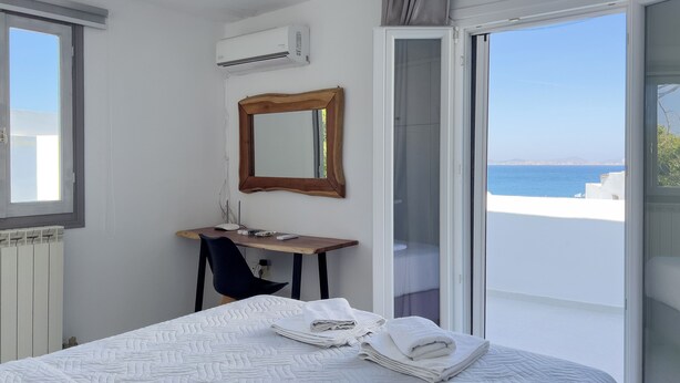 A bedroom with a bed, a desk, a chair, a mirror, and a view of the sea. - Naxian Downtown Residence (Naxos Chora)