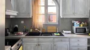 A kitchen with grey cabinets, a window with a sheer curtain, a sink, a stove, and various appliances. - Naxian Downtown Residence (Naxos Chora)