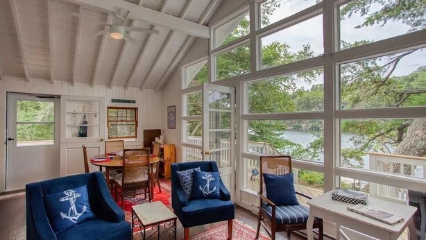 A spacious room with large windows overlooking a lake, furnished with blue chairs and a wooden table. - Charming Cottage on the Great Pond in Boothbay Harbor, Maine (Boothbay Harbor)