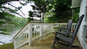 A wooden deck with a white railing, a rocking chair, and a view of a lake surrounded by trees. - Charming Cottage on the Great Pond in Boothbay Harbor, Maine (Boothbay Harbor)
