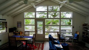 A spacious room with a high ceiling, large windows offering a view of the outdoors, a dining area with a table and chairs, and a seating area with sofas. - Charming Cottage on the Great Pond in Boothbay Harbor, Maine (Boothbay Harbor)