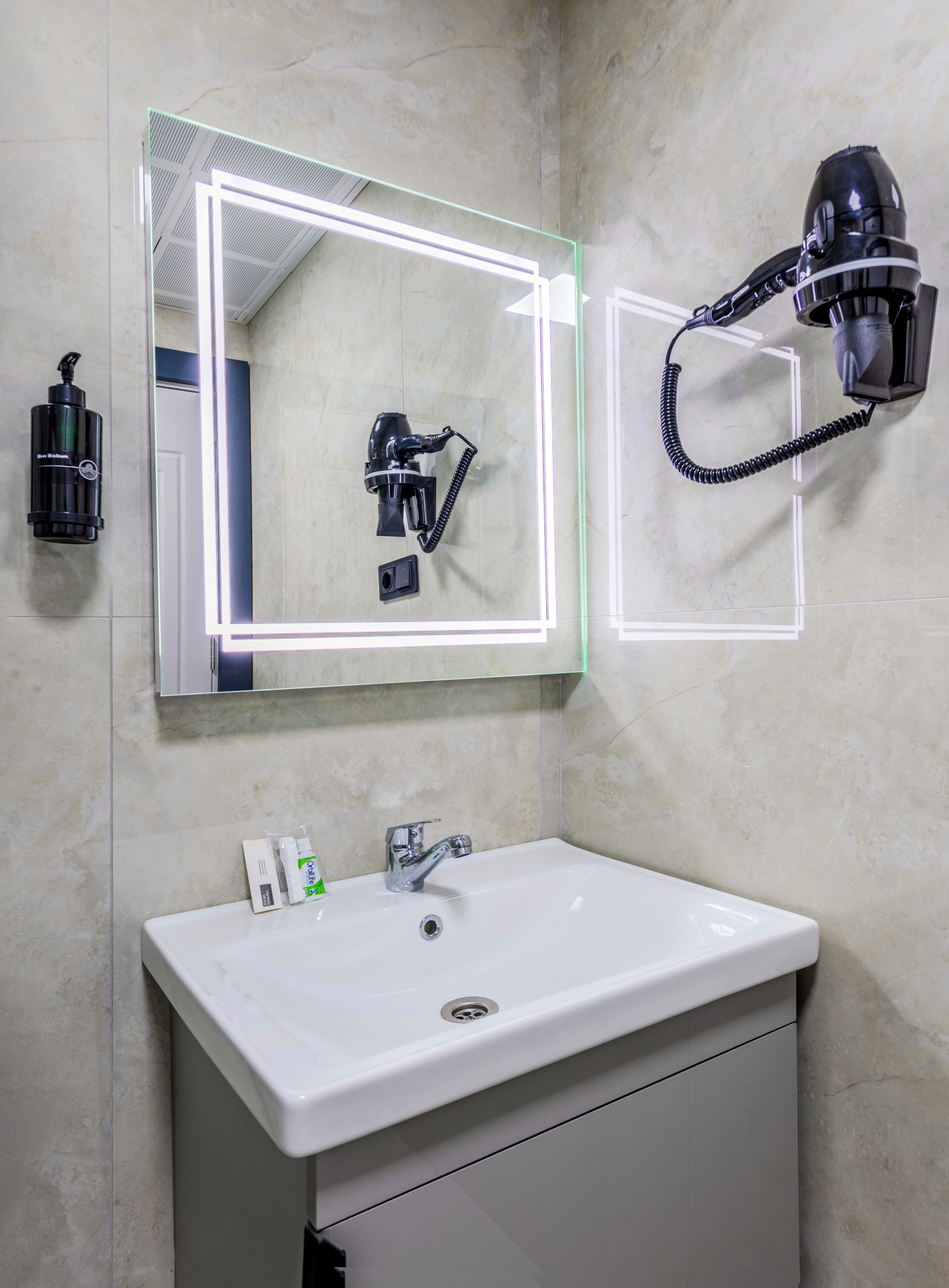Une salle de bain avec un lavabo blanc, un miroir avec éclairage à DEL, un sèche-cheveux et une bouteille de shampoing.
