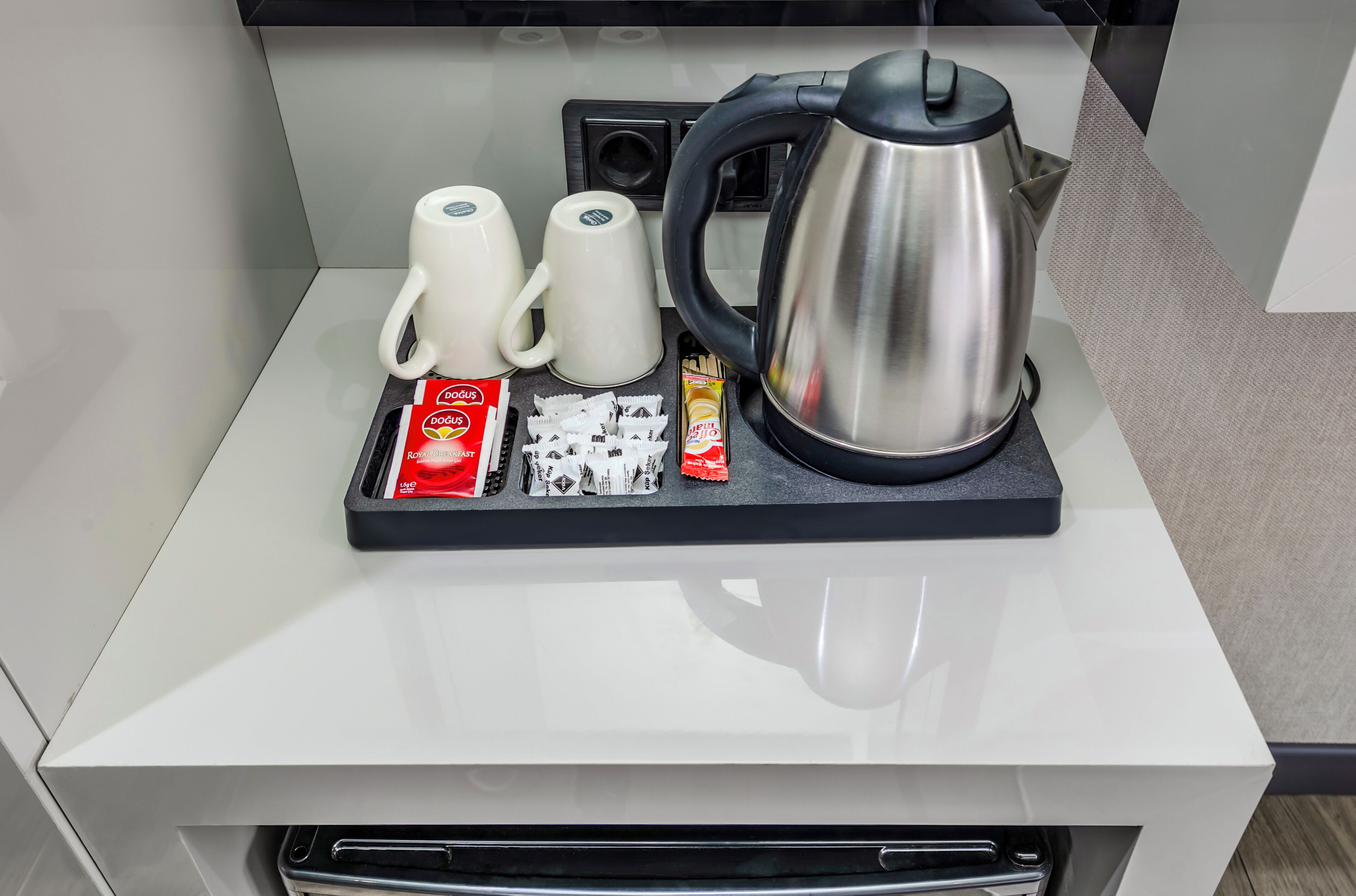 Ein Kaffee-Bereich mit Wasserkocher, zwei Tassen, einem Tablett mit Kaffeekapseln, einer Packung Instantkaffee und Zucker.