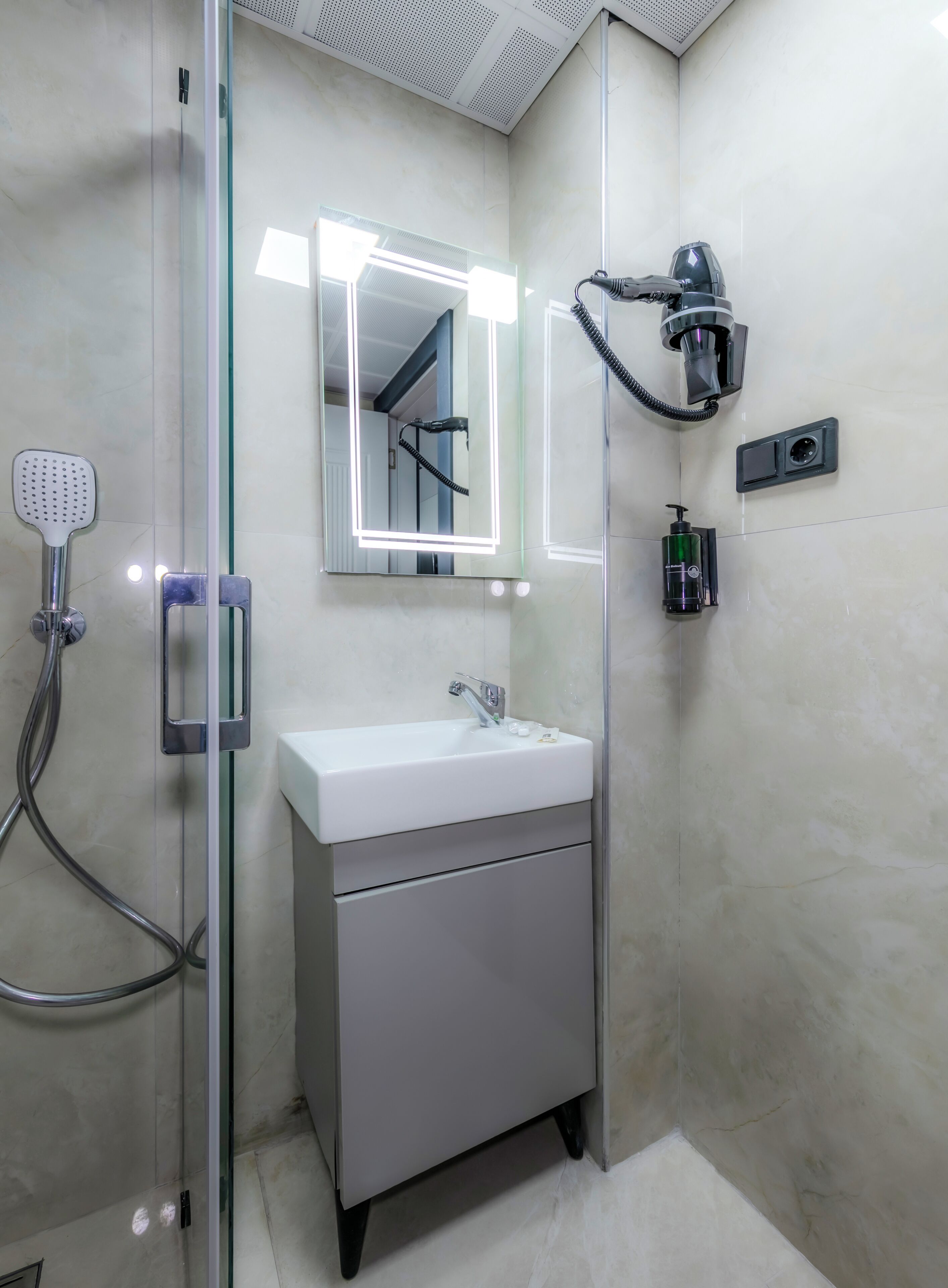 A modern bathroom with a glass-enclosed shower, a white sink, and a hairdryer mounted on the wall.