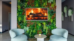 A room with a wall-mounted TV displaying a fireplace, surrounded by lush artificial foliage. There are two light blue armchairs and a small round table in the foreground. - Dedem Hotel Premium Çorlu (Corlu)