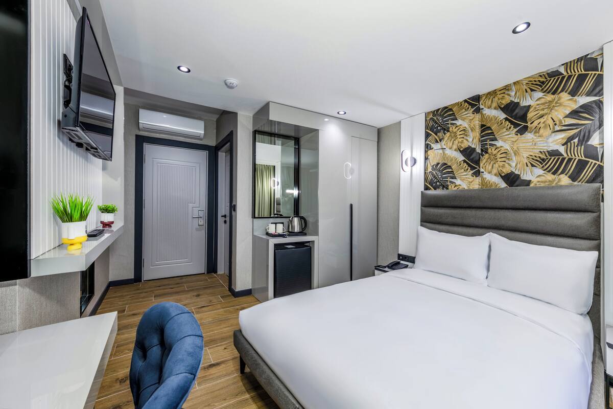 A modern hotel room with a large bed, a desk with a chair, a small kitchenette, and a TV mounted on the wall.