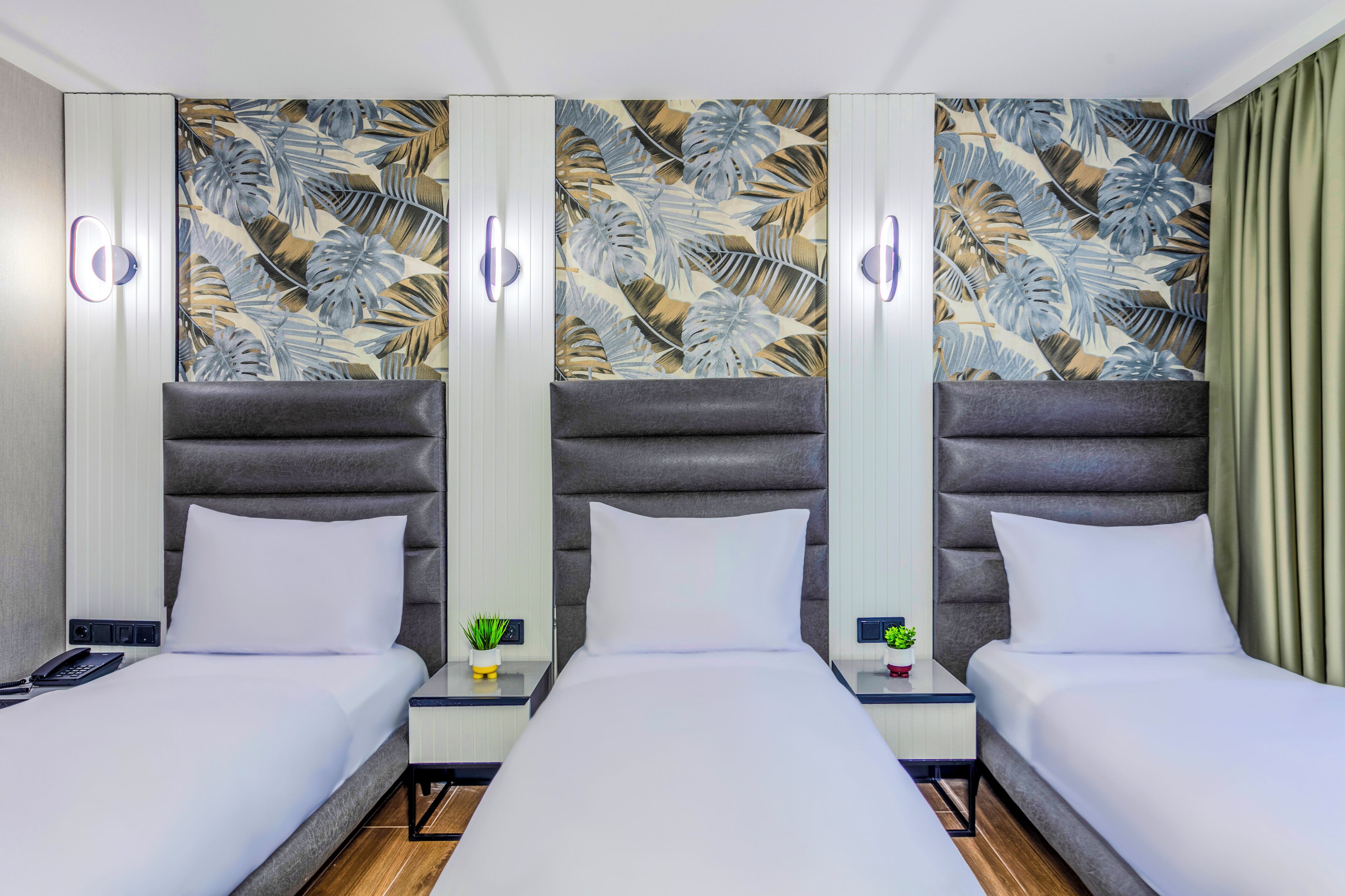 Habitación de hotel con tres camas, pared con estampado de hojas tropicales y mesitas de noche con pequeñas plantas.