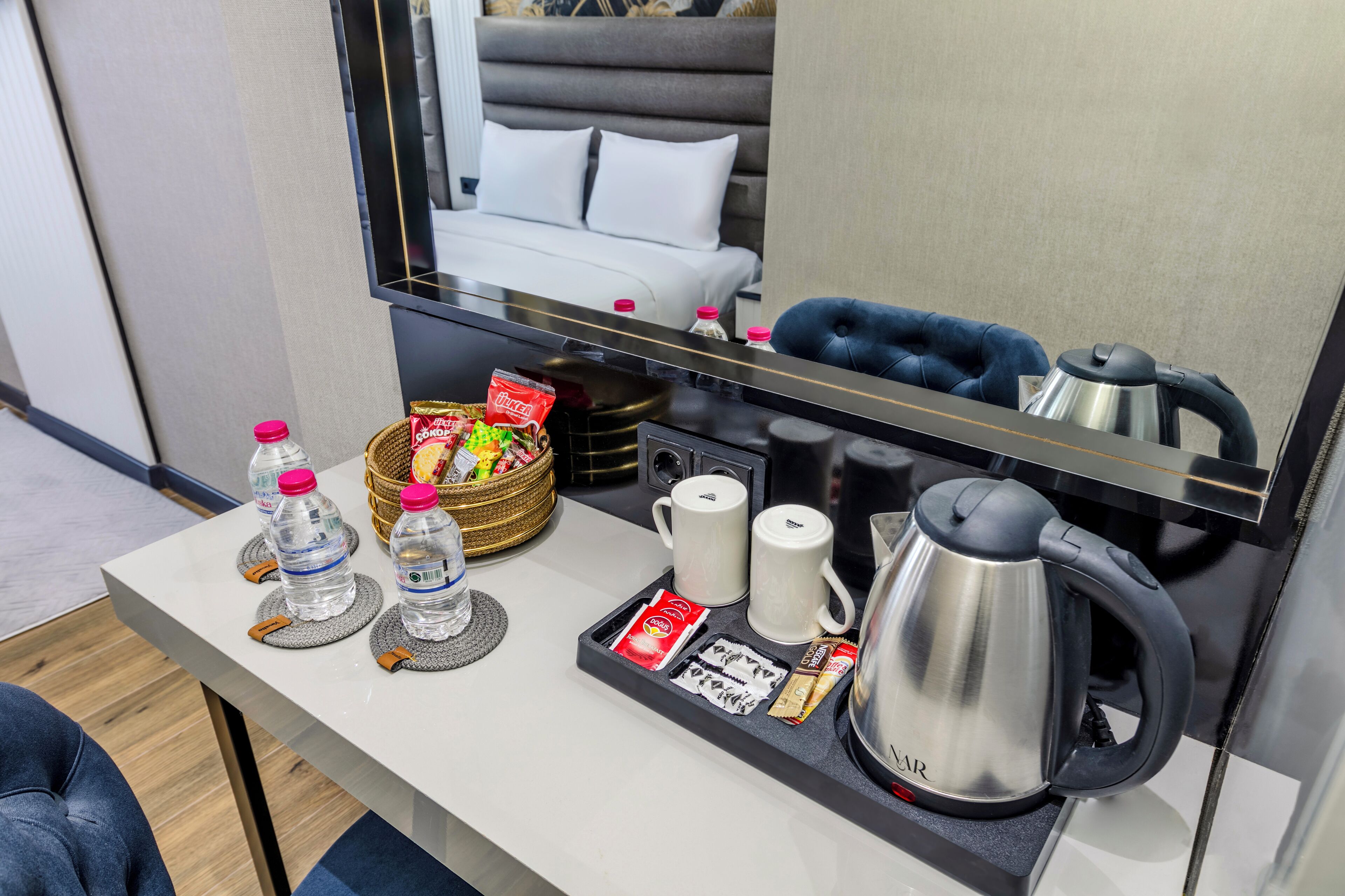 Habitación de hotel con una bandeja de desayuno ordenada que incluye botellas de agua, una cesta de aperitivos, tazas de café y una tetera.