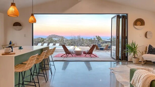 A modern kitchen with a bar area, a dining space with chairs, and a view of a mountainous landscape through large glass doors. - Luxury Spa home w/ Entertainers Pool, hot tub & fire pits! (Yucca Valley)