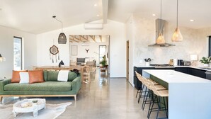 A spacious living area with a green sofa, a dining table, and a kitchen with a large island and bar stools. - Luxury Spa home w/ Entertainers Pool, hot tub & fire pits! (Yucca Valley)