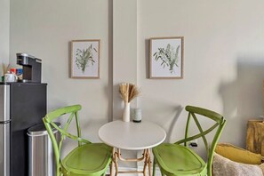 A compact kitchen with a refrigerator, a sink, and a small dining table with two chairs. There are framed botanical prints on the wall and a vase with dried plants on the table.
